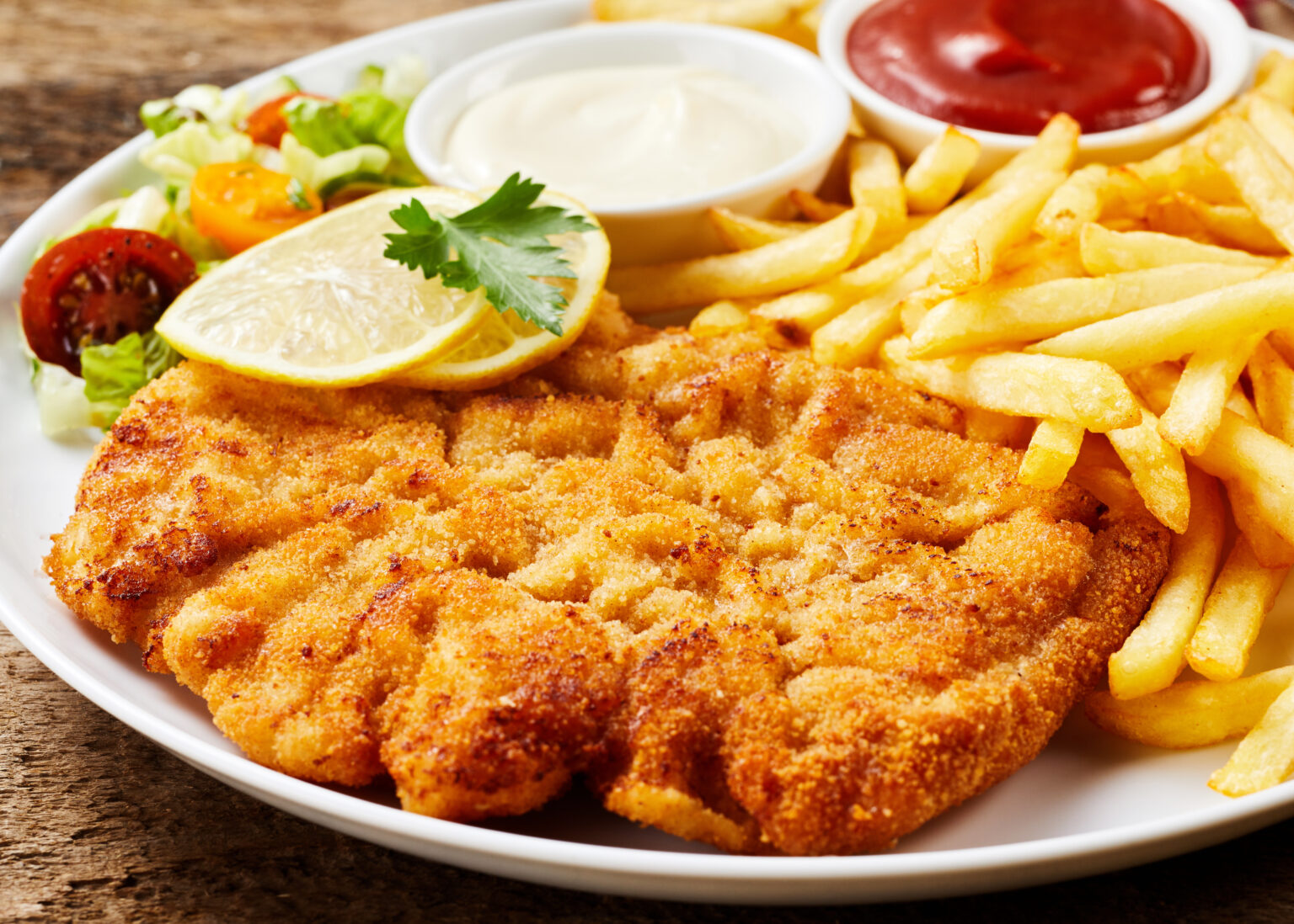 Schnitzel and French fries dish served with ketchup and mayonnaise, lemon and vegetables, close-up on white plate